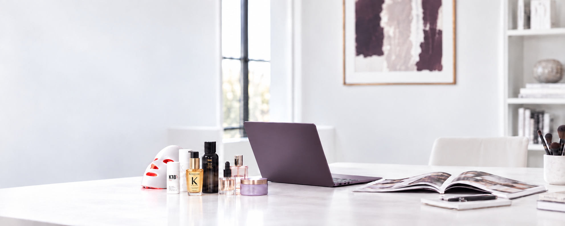 Luxury beauty editor desk with a purple laptop, open magazine, hair products, and a red light therapy mask.